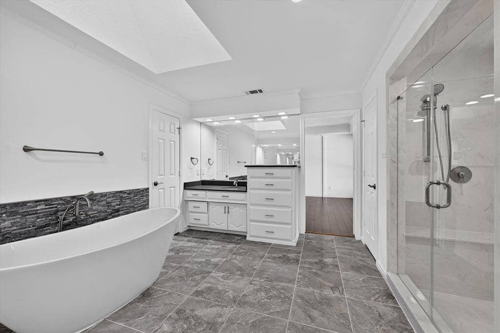 Freestanding soaking tub with a stacked stone tile accent wall, glass-enclosed shower, dual vanity with undermount sinks, and stone countertop