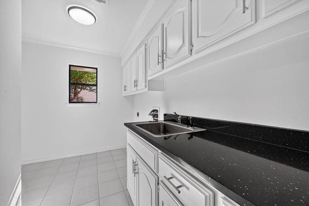 Built-in cabinetry with white paneling and silver-tone hardware, black speckled countertop, stainless steel sink, tiled flooring, and a window providing natural light, much cabinet space, extra room for freezer