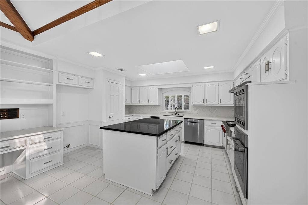 White cabinetry, stainless steel appliances, central island with dark countertop, tile flooring, and built-in desk with shelving