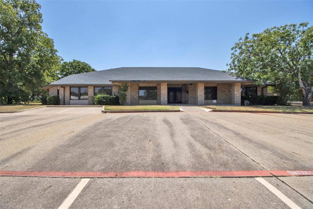 3210 W Northwest Loop 323, Tyler, TX 75702 Brick exterior with a dark shingle roof, multiple windows, established perimeter landscaping, and an expansive paved parking area