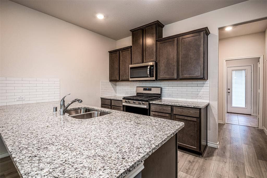 2721 Franklin Drive, Celina, TX 75009 Kitchen featuring granite countertops, stainless steel appliances, dark wood cabinetry, a subway tile backsplash, and wood-finish flooring