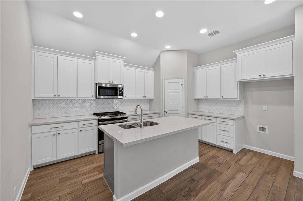 5308 Tupper Avenue, Fate, TX 75087 Kitchen featuring white cabinetry, a central island with an undermount sink, stainless steel range and microwave, tiled backsplash, and wood-finish flooring