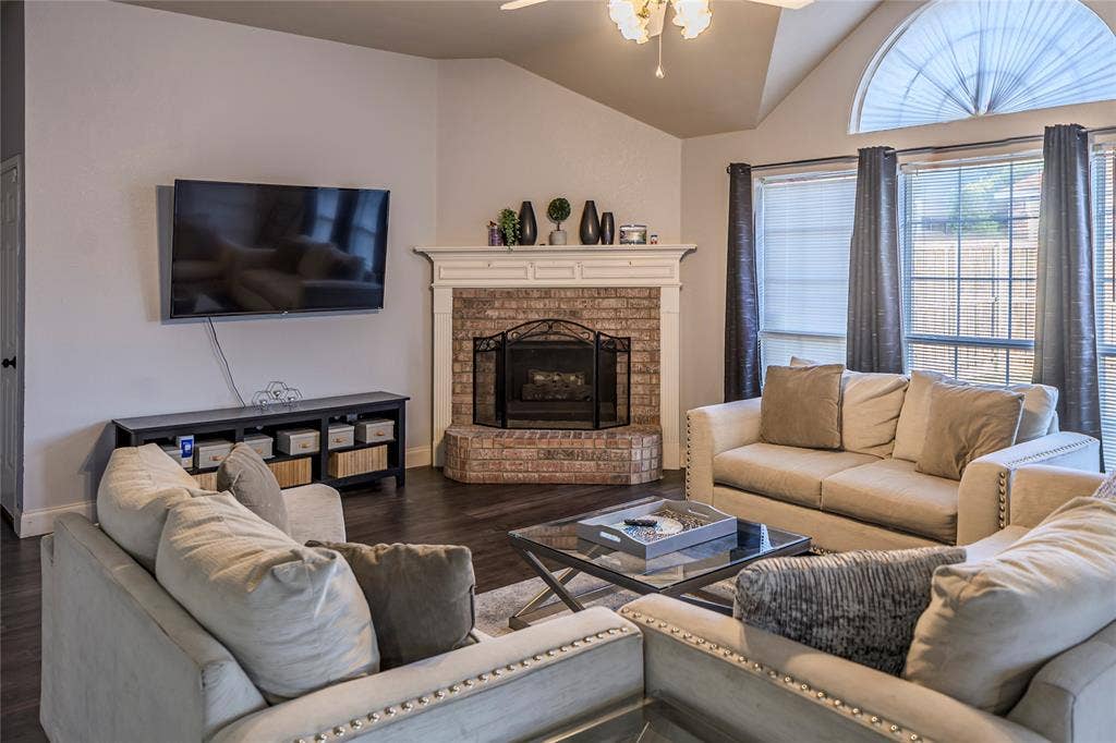 Vaulted ceiling, brick fireplace with mantel, dark wood-finish flooring, large arched window, and ceiling fan with light fixture