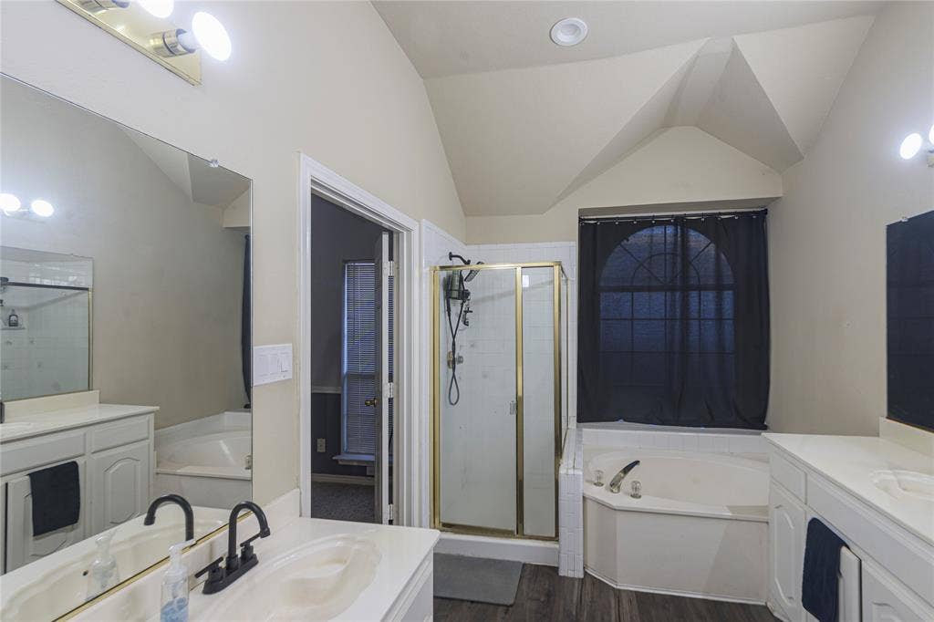 Dual vanities with white countertops, corner soaking tub, separate shower enclosure with gold-tone framing, wood-finish flooring, and vaulted ceiling