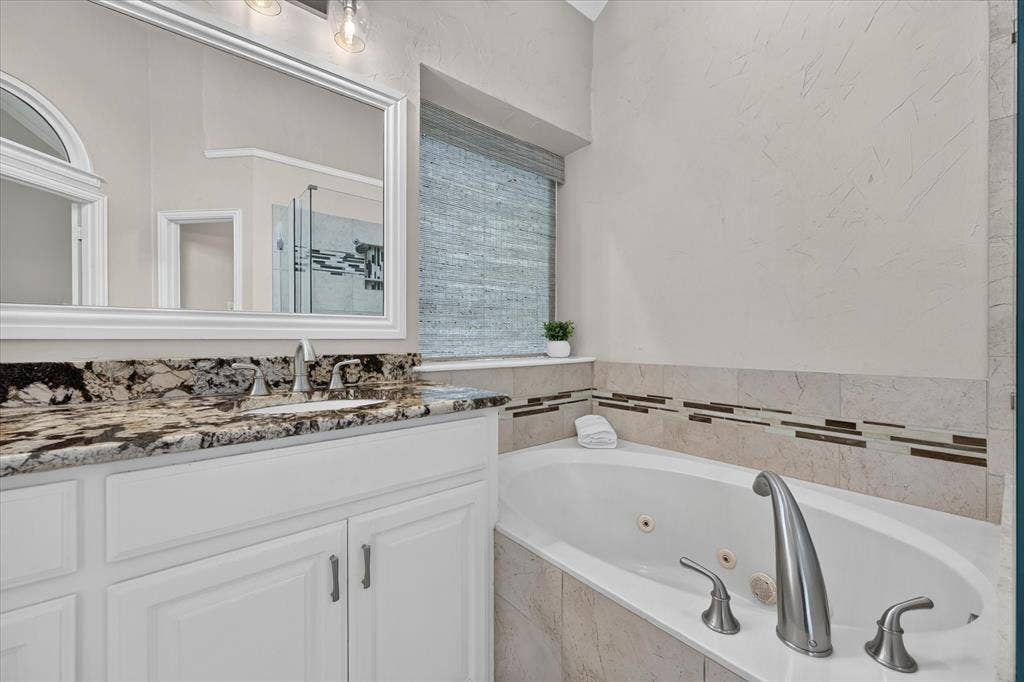 10307 Perkins Drive, Irving, TX 75063 White vanity with a stone countertop and undermount sink, framed mirror, and a built-in jetted tub with tile surround and decorative accent band