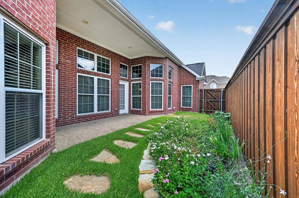 10307 Perkins Drive, Irving, TX 75063 Red brick exterior with multiple windows, covered patio, stone paver pathway, planted garden bed, and wood privacy fencing