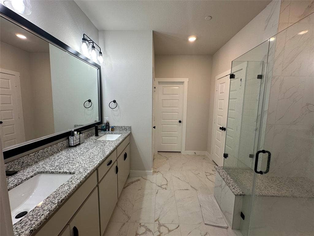 Spa-inspired primary bathroom featuring dual vanities with speckled quartz countertops, a frameless glass-enclosed shower with marble-look tile surround, and recessed ceiling lighting