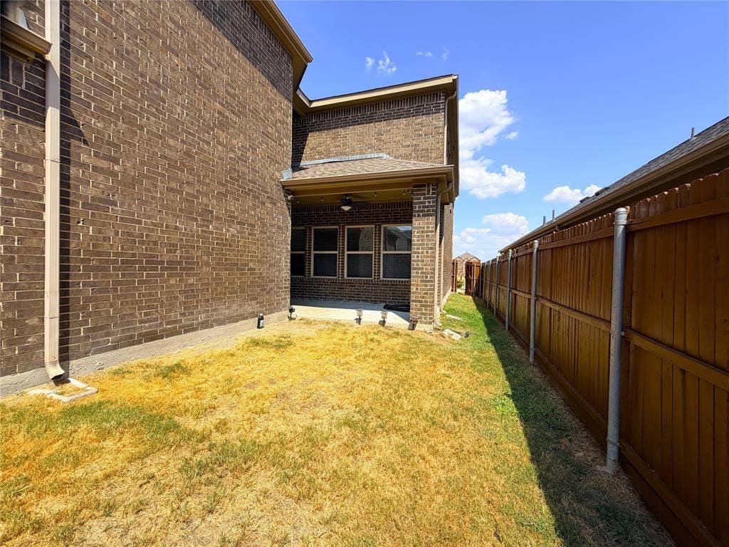 4103 Portrush Drive, Heartland, TX 75126 Spacious backyard with level lawn and wooden privacy fencing, adjoining a brick residence featuring a covered patio entry and oversized transom window beneath clear blue skies