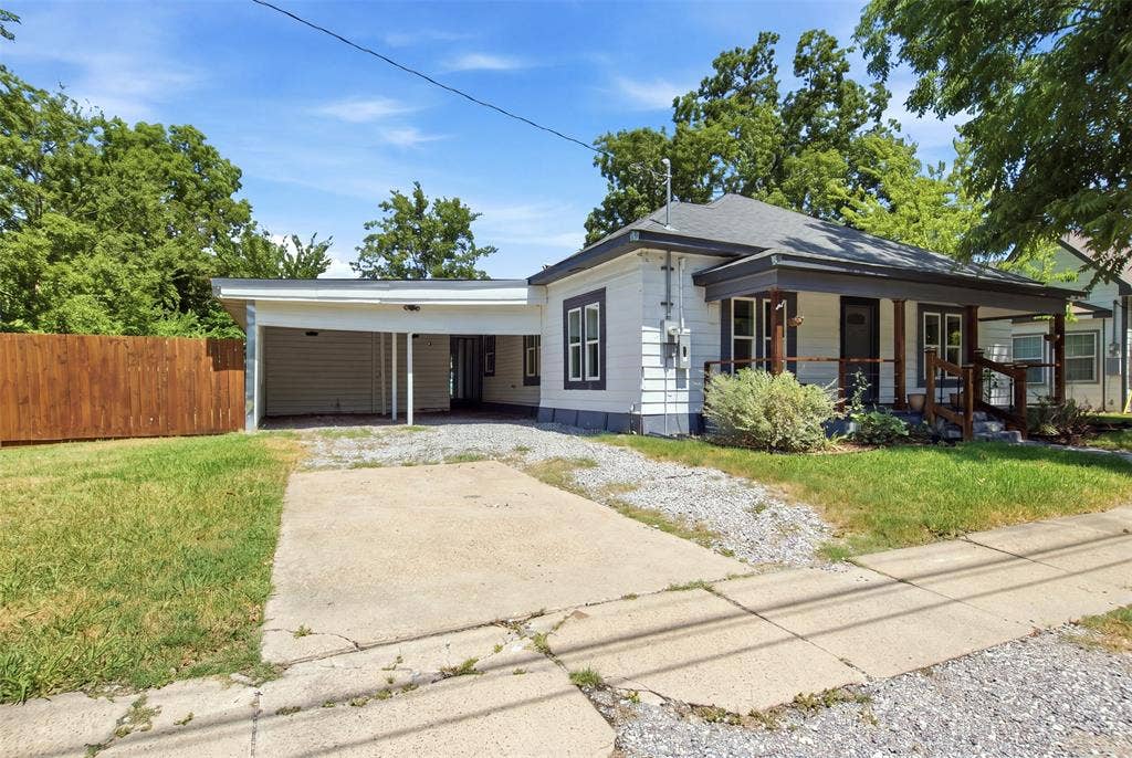 1812 Oneal Street, Greenville, TX 75401 Classic single-story home featuring a front-facing carport with overhead door, mature landscaping with established trees, concrete driveway, and a covered front porch entry with board-and-batten siding and traditional window detailing