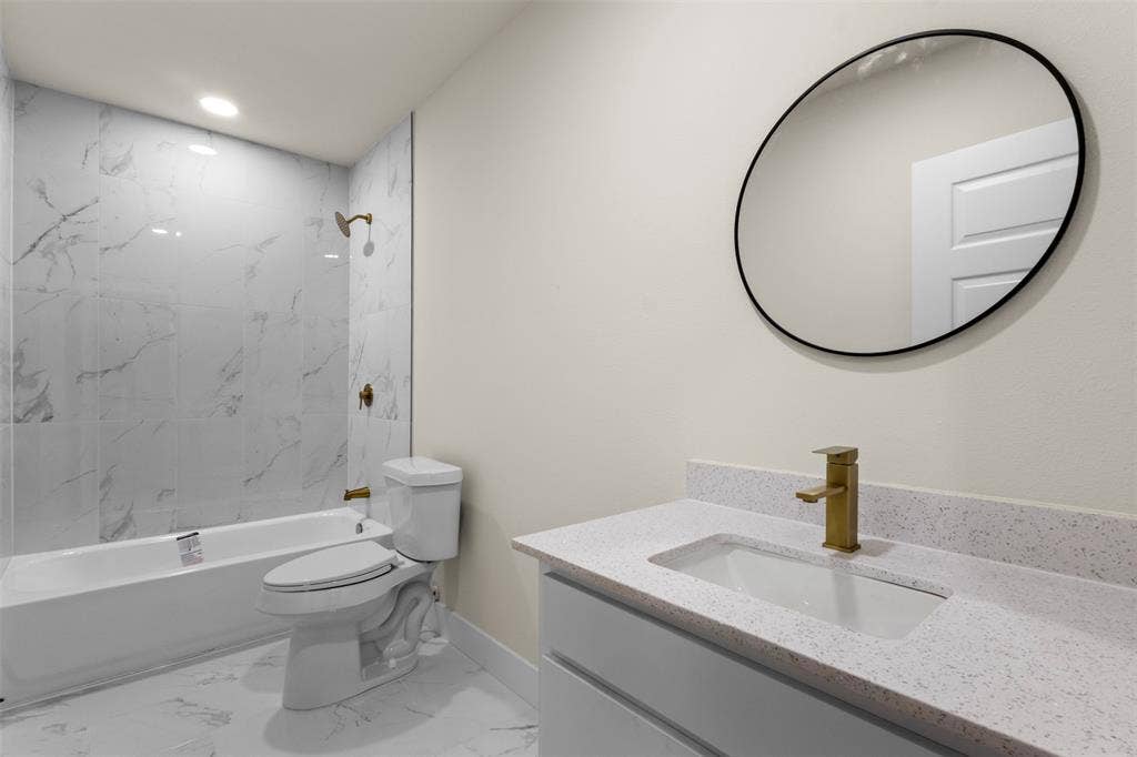212 E Marguerita Drive, Garland, TX 75040 Marble-look tile flooring, white bathtub with a bronze shower fixture, white vanity with a bronze faucet, white countertop with integrated sink, and a circular mirror with a black frame