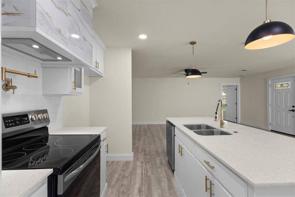 Kitchen featuring white cabinetry with gold-toned hardware, quartz countertops, a central island with an undermount double-basin sink, wall-mounted pot filler, and wood-finish flooring