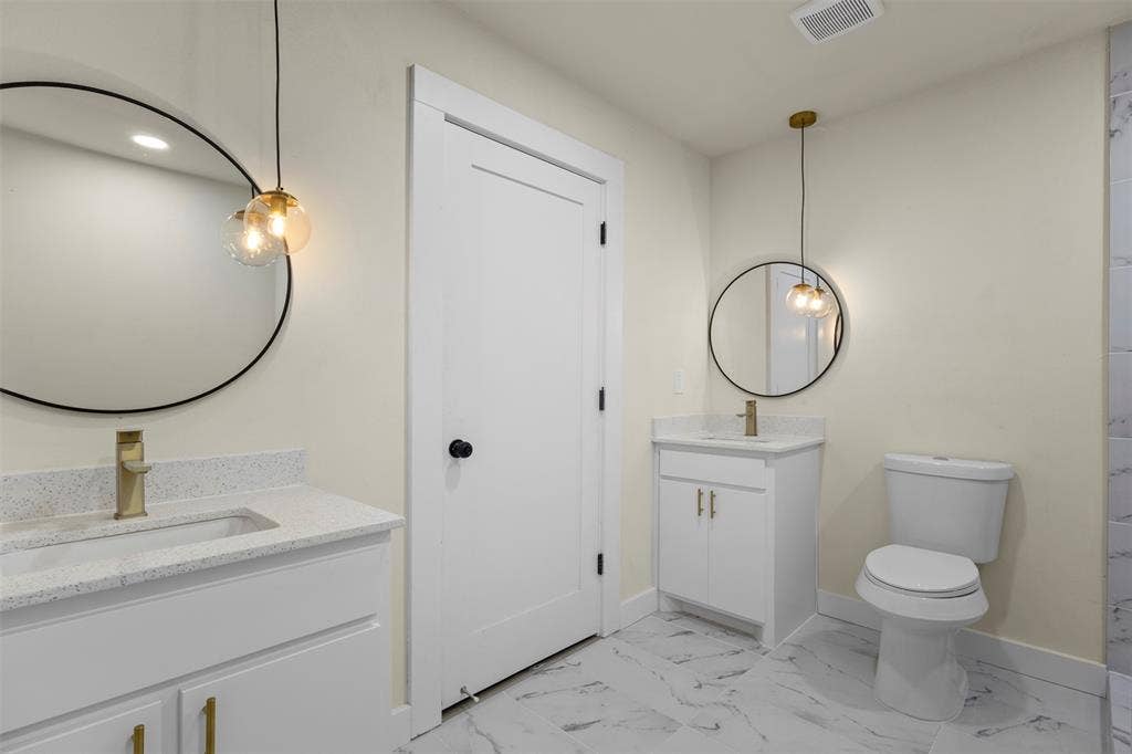 Double vanity bathroom featuring white cabinetry with gold-toned hardware, quartz-composite countertops, round wall-mounted mirrors, pendant lighting, and stone-finish tile flooring