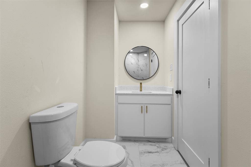 Modern vanity with white cabinetry, stone-finish countertop, undermount sink, and brass-tone hardware, complemented by a round wall-mounted mirror, brass-tone faucet, and stone-patterned tile flooring
