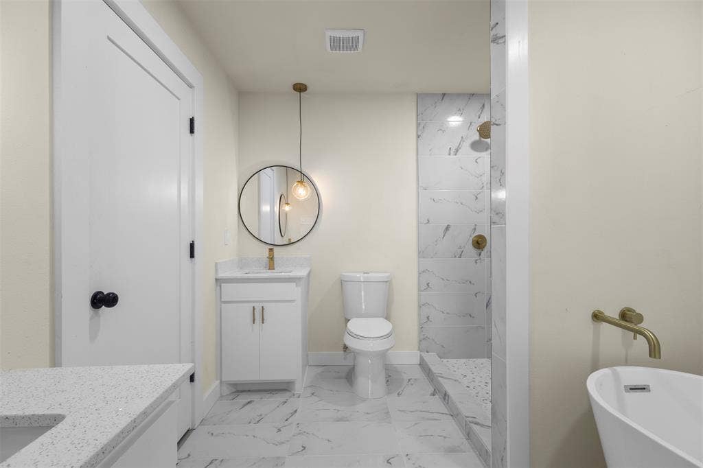 White vanity cabinet with quartz countertop, circular wall-mounted mirror, brass-finish fixtures, stone-patterned tile flooring, and a walk-in shower with stone-patterned tile surround