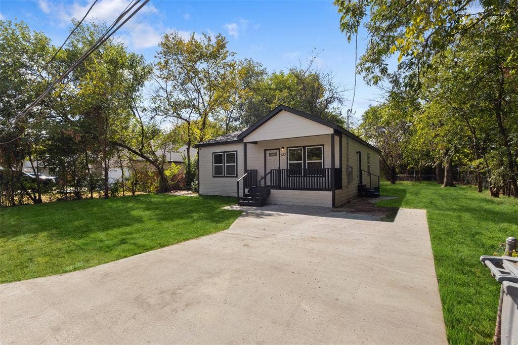 333 Levant Avenue, Dallas, TX 75217 Contemporary exterior with light siding and dark trim, featuring a covered front porch with dark railing, a concrete driveway, and surrounding mature trees