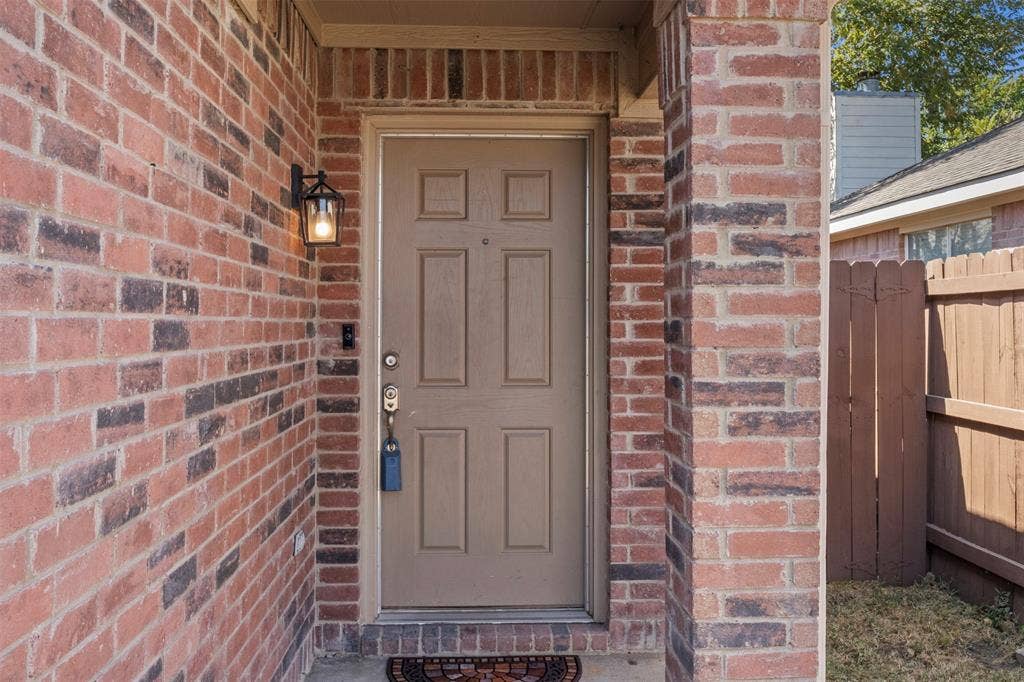 Brick exterior facade with a paneled entry door, exterior wall lantern, and partial wood privacy fence
