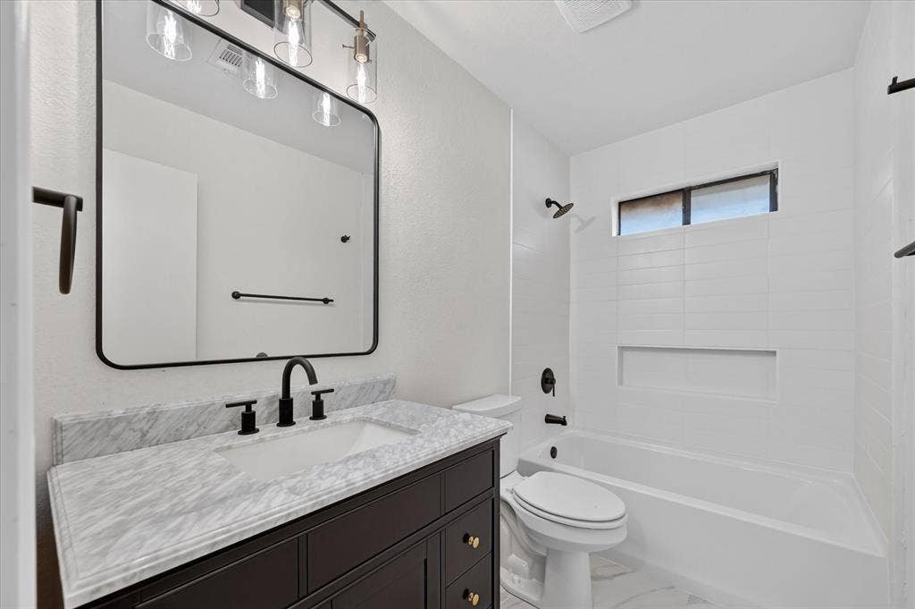 1528 Estrella Drive, Plano, TX 75074 Rectangular vanity mirror with integrated lighting, dark wood vanity with gold-tone hardware, undermount sink with matte black fixtures, white tiled shower surround with built-in niche, and a window above the shower