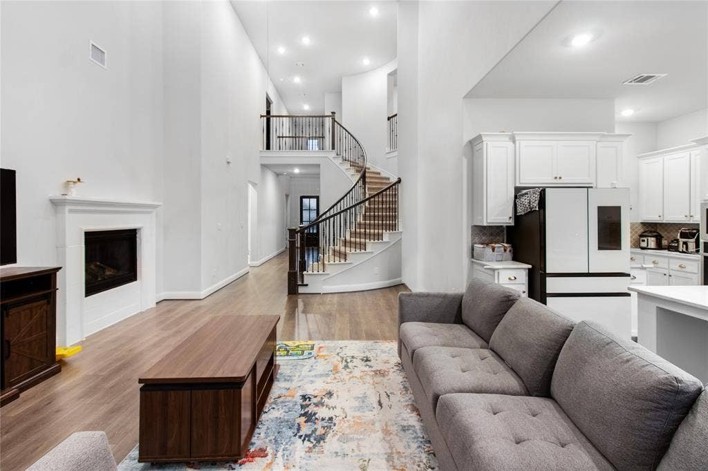 1531 20th street, Argyle, TX 76226 Open-concept living space featuring soaring ceilings with recessed lighting, a curved staircase with metal railings and wood treads, hardwood flooring throughout, a gas fireplace with white surround, and a modern kitchen with white cabinetry and stainless steel appliances