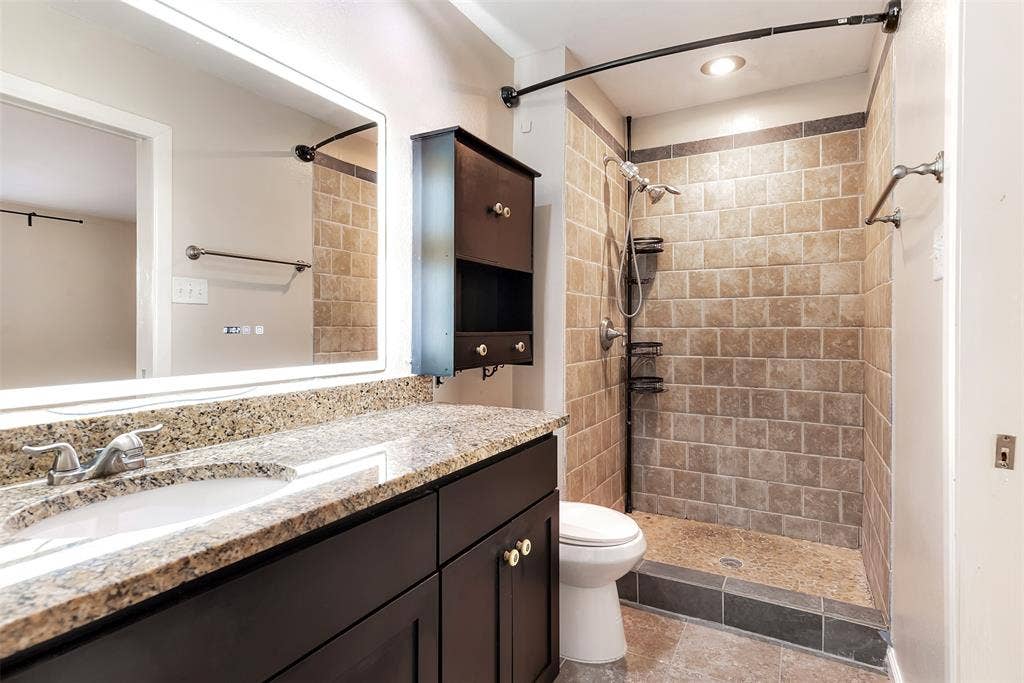 925 Lombardy Drive, Plano, TX 75023 Walk-in shower with natural stone-look tiling, granite-look countertop, dark wood-finish vanity, and illuminated mirror