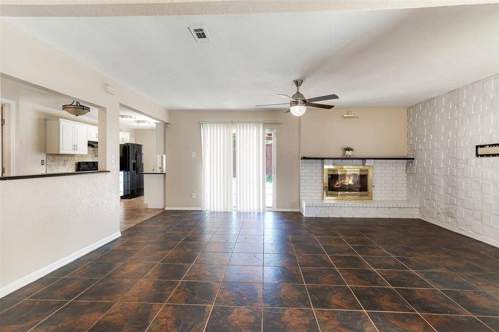 925 Lombardy Drive, Plano, TX 75023 Tile flooring, brick fireplace with a brass-finish insert, white accent wall, ceiling fan, and sliding glass door with vertical blinds