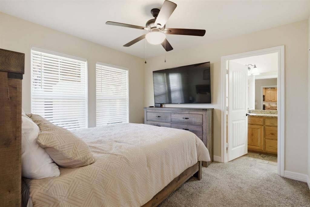 213 Freeman Street, Lindale, TX 75771 Carpeted bedroom with a ceiling fan, two windows with blinds, and an open doorway revealing a bathroom with wood cabinetry and a stone countertop
