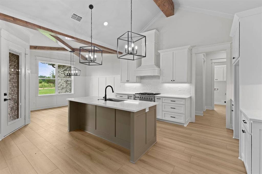 13914 Cedar Hill Lane, Tyler, TX 75704 Vaulted ceiling with exposed wood beams, wood-finish flooring, central kitchen island with integrated sink, white shaker-style cabinetry, and stainless steel range with custom hood