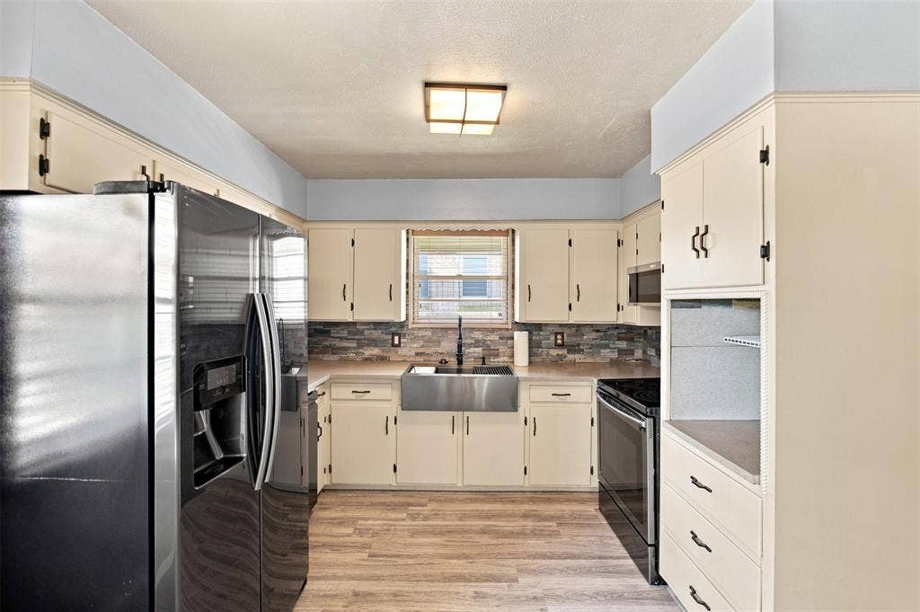 2022 Normandy Drive, Irving, TX 75060 Kitchen featuring wood-finish flooring, a stainless steel farmhouse sink, stacked stone backsplash, and cream cabinetry with dark hardware