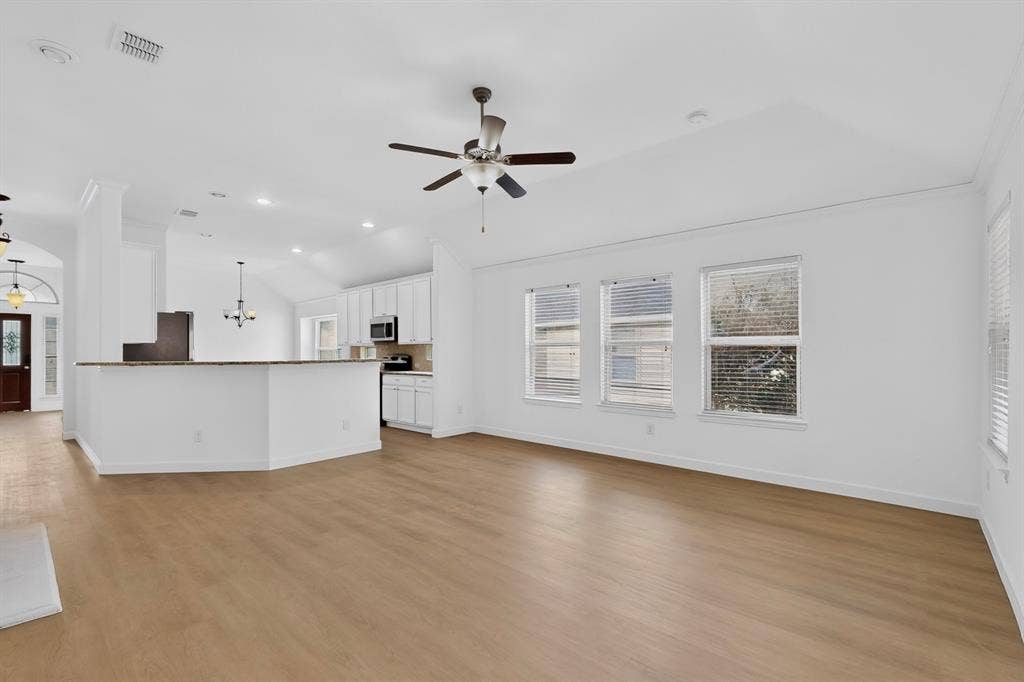 Open-concept interior featuring vaulted ceilings, wood-finish flooring, a kitchen breakfast bar, and multiple windows with horizontal blinds