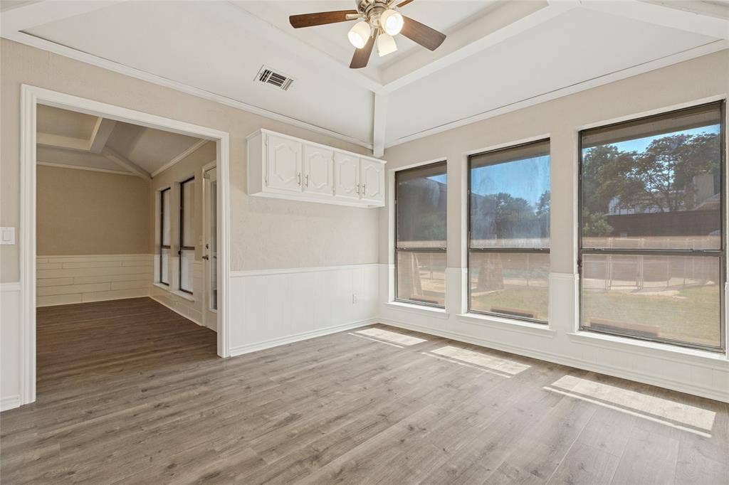 5013 Country Place Drive, Plano, TX 75023 Vaulted ceiling with a ceiling fan, wood-finish flooring, three large windows, white cabinetry, and wainscoting