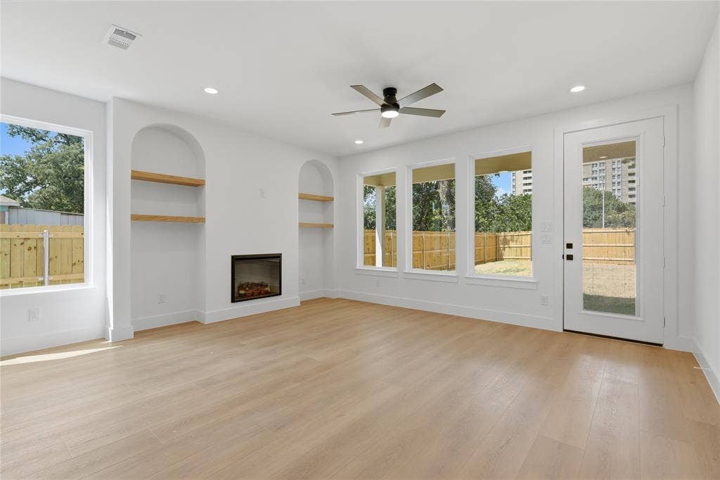 3300 S Harwood Street, Dallas, TX 75215 Spacious living area featuring wood-finish flooring, an electric fireplace, two arched built-in shelving units, a ceiling fan with integrated lighting, and multiple windows and a glass-paneled door opening to the exterior