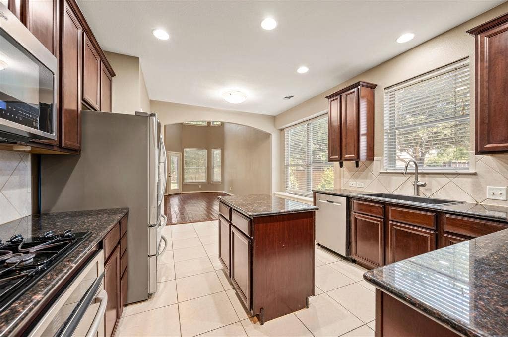6201 Crator Drive, McKinney, TX 75070 Kitchen featuring dark wood cabinetry, stainless steel appliances, granite countertops, and tile flooring that has an open concept to the living room. Also love the window over the sink!