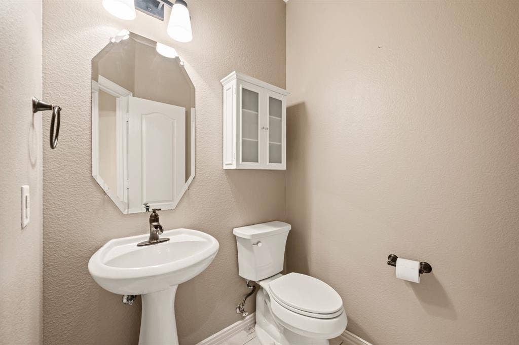 6201 Crator Drive, McKinney, TX 75070 Half bathroom downstairs with Pedestal sink with a bronze-finish faucet, octagonal mirror, two-light vanity fixture, white wall-mounted cabinet, and toilet