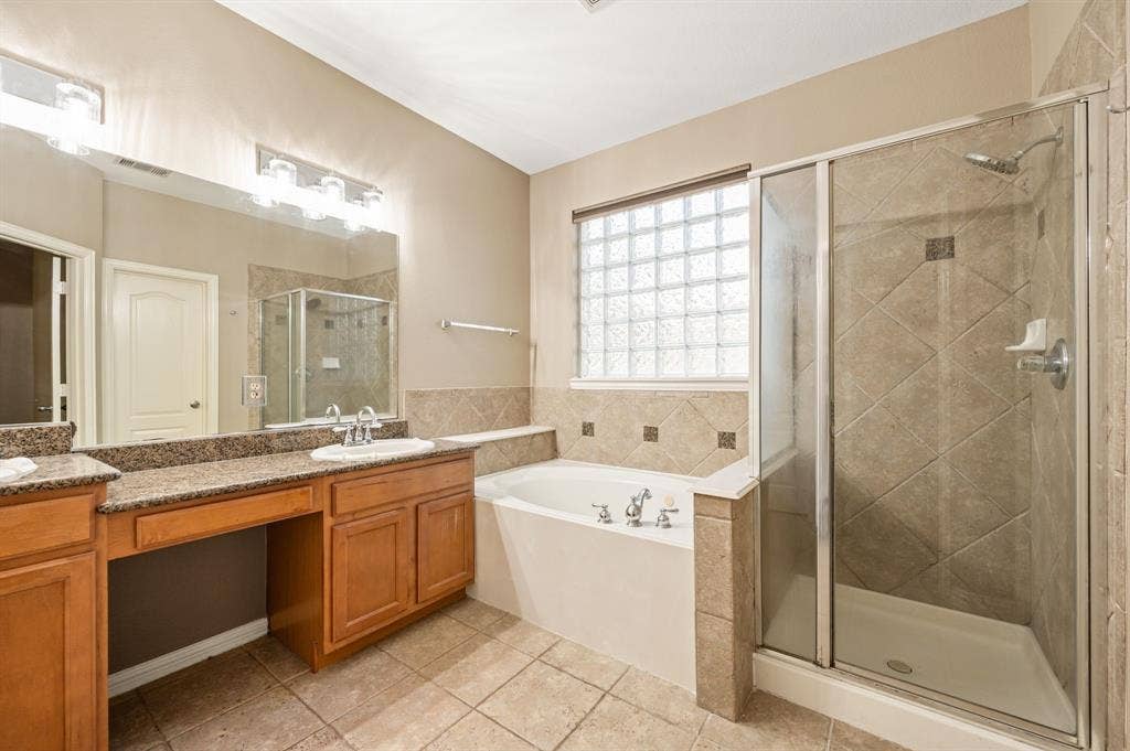 6201 Crator Drive, McKinney, TX 75070 Bathroom featuring a built-in soaking tub, glass-enclosed shower with tile surround, vanity with dual sinks and granite countertop, decorative glass block window and updated lighting