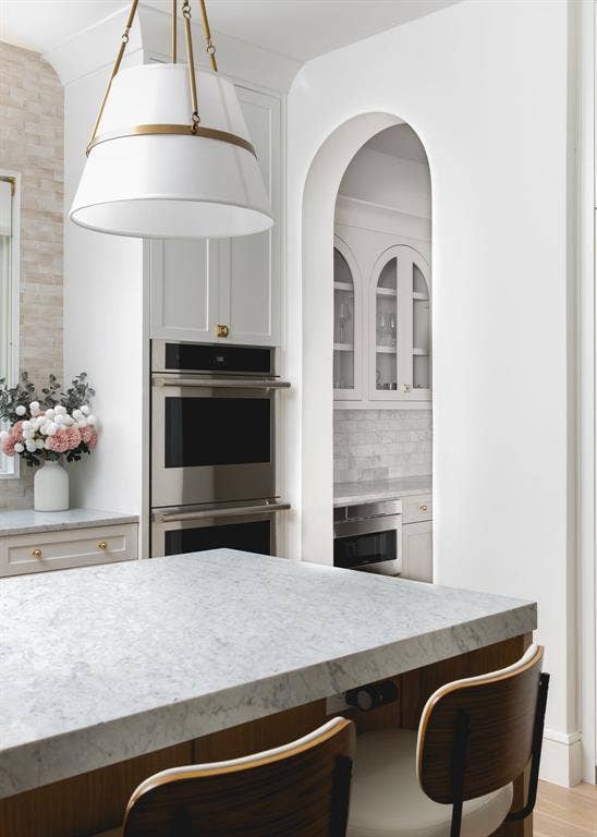 Kitchen featuring a light-toned stone countertop, integrated stainless steel double ovens, and a decorative pendant light