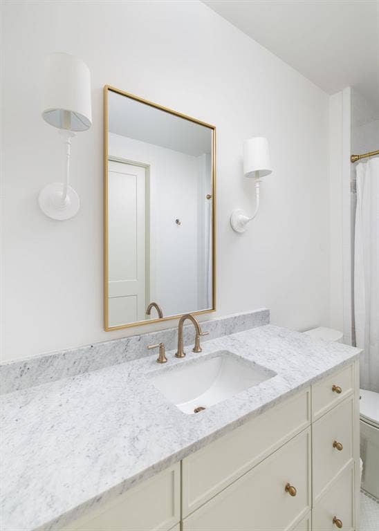White vanity with integrated sink, stone countertop, gold-framed mirror, and brass-finish fixtures
