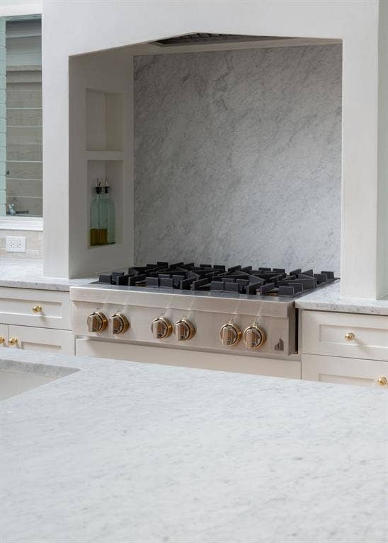 Built-in gas range with gold-tone hardware, integrated range hood, stone slab backsplash, stone countertops, and recessed shelving