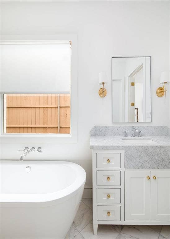 Freestanding soaking tub with wall-mounted faucet, single vanity with white cabinetry, stone countertop, undermount sink, and wall-mounted sconces