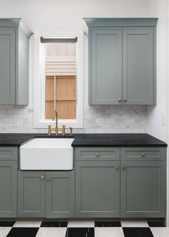 Farmhouse sink with a gold-tone bridge faucet, dark countertops, gray shaker cabinetry, and a white subway tile backsplash