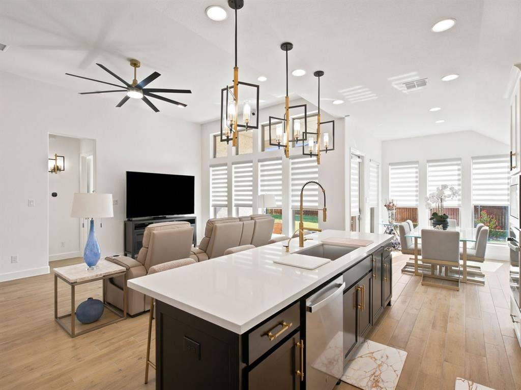 Open-concept living space with wood-finish flooring, a kitchen island with a sink and gold-tone faucet, and multiple windows featuring horizontal blinds