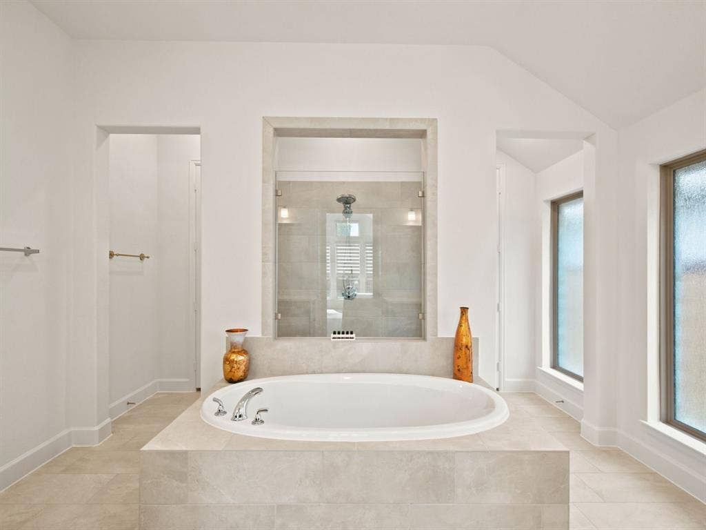 Deep soaking tub with chrome fixtures, tiled surround, glass-enclosed shower, and opaque window panels