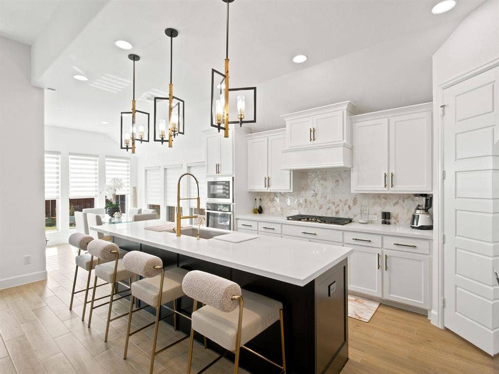 Chef's kitchen featuring white cabinetry with brushed gold hardware, a black waterfall-edge island with seating for multiple guests, quartz countertops, stainless steel gas cooktop, pendant lighting with geometric frames, and a subway tile backsplash