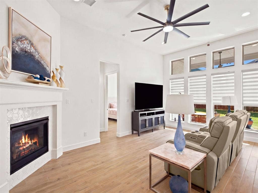 Light wood-finish flooring, a fireplace with a white mantel and hexagonal tile surround, an angular ceiling fan, and multiple windows with blinds