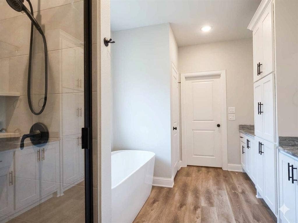 11034 Oak Springs Cove, Tyler, TX 75703 Freestanding white soaking tub, wood-finish flooring, white cabinetry with dark hardware, glass-enclosed shower with dark fixtures, and light-toned countertops