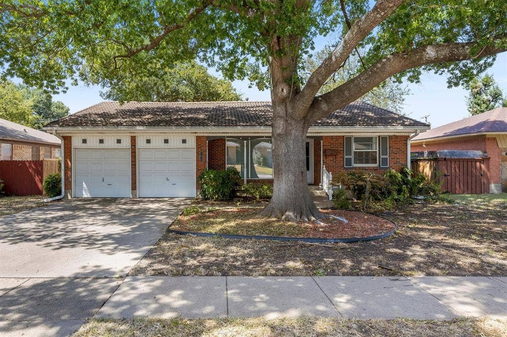 1013 Bandera Lane, Garland, TX 75040 Brick exterior featuring a two-car garage, front landscaping, and a mature tree