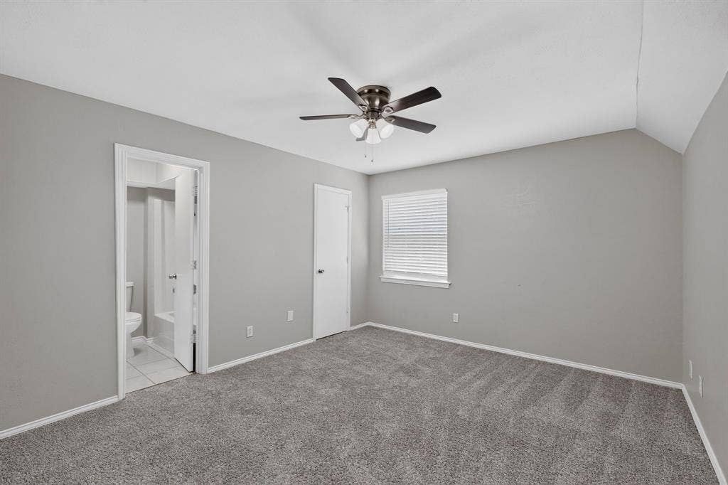 Carpeted flooring, vaulted ceiling, ceiling fan, window with blinds, and an attached bathroom with a toilet and tub