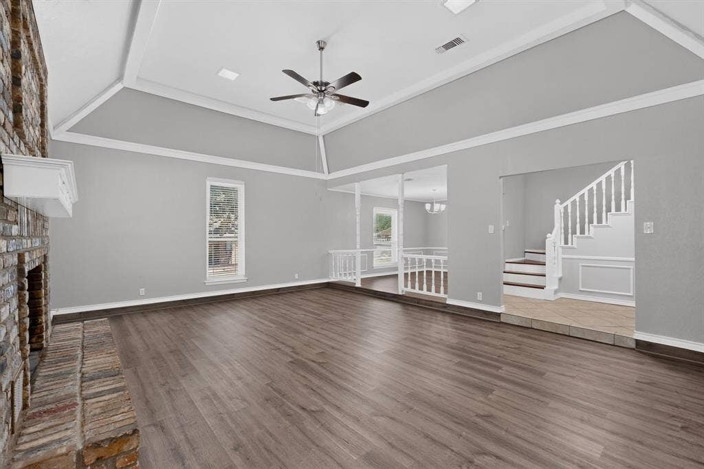 Spacious room featuring vaulted ceilings, a prominent brick fireplace with a white mantel, wood-finish flooring, and a staircase with white balusters