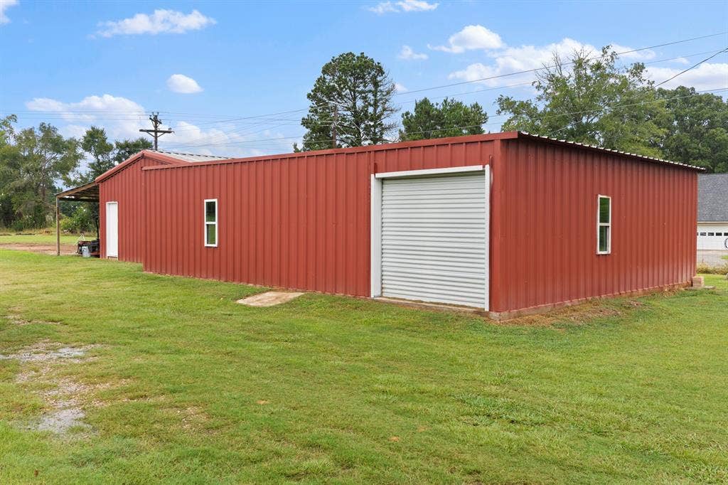 22477 County Road 2166, Troup, TX 75789 Red-colored metal building featuring a corrugated metal exterior, a roll-up garage door, white framed windows, a white entry door, and an attached covered extension