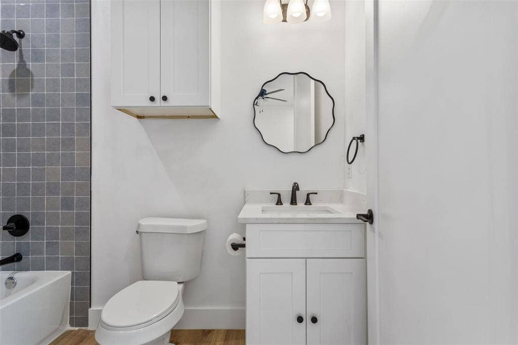 Modern bathroom featuring a white vanity with dual cabinet doors and an integrated sink, matte black hardware and faucet, a scalloped-edge mirror with black frame, white subway tile wall treatment, wood-finish flooring, and a white toilet with elongated bowl