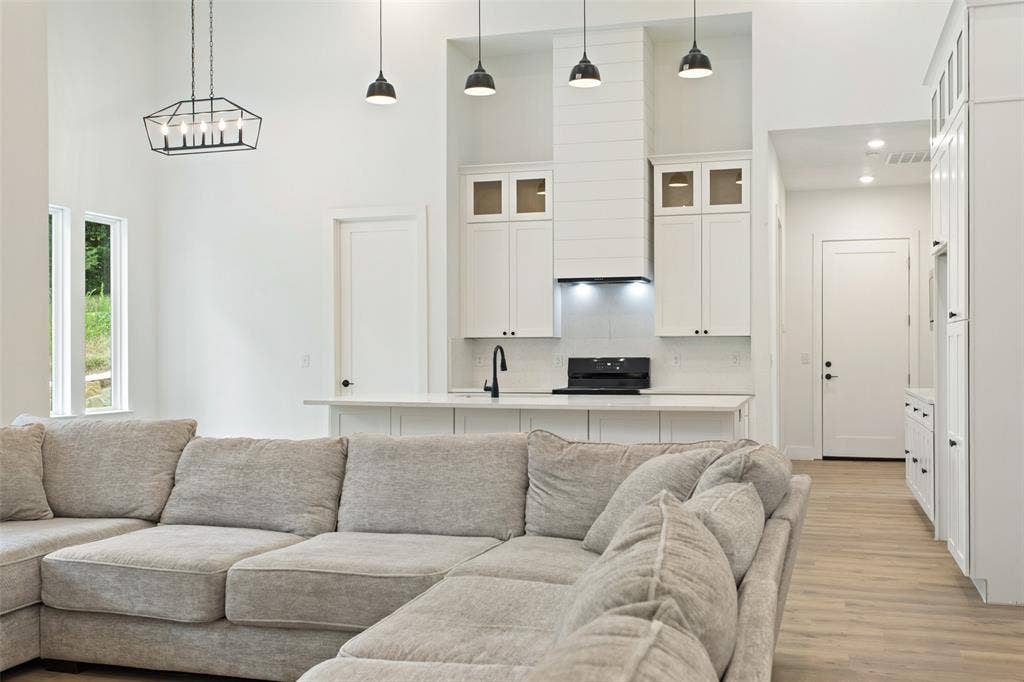 Open-concept living and kitchen area featuring a sectional seating arrangement, light wood-finish flooring throughout, custom white cabinetry with glass-front upper storage, stainless steel appliances, pendant lighting, and abundant natural light from large windows