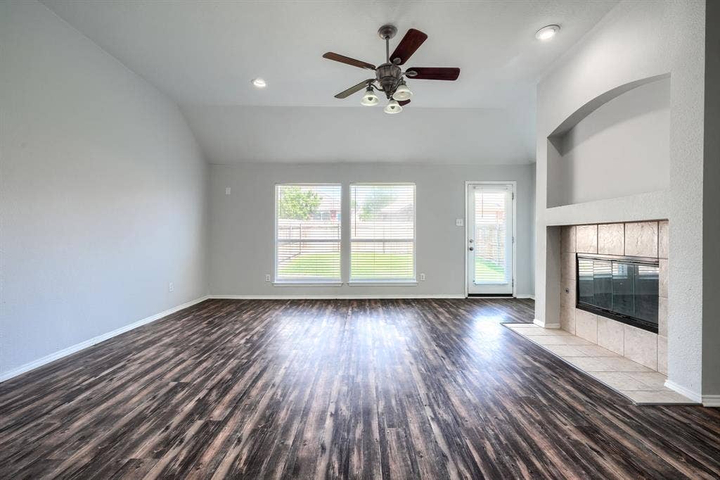 Vaulted ceiling with a ceiling fan, wood-finish flooring, two large windows, a glass-paned door, and a fireplace with a tiled surround